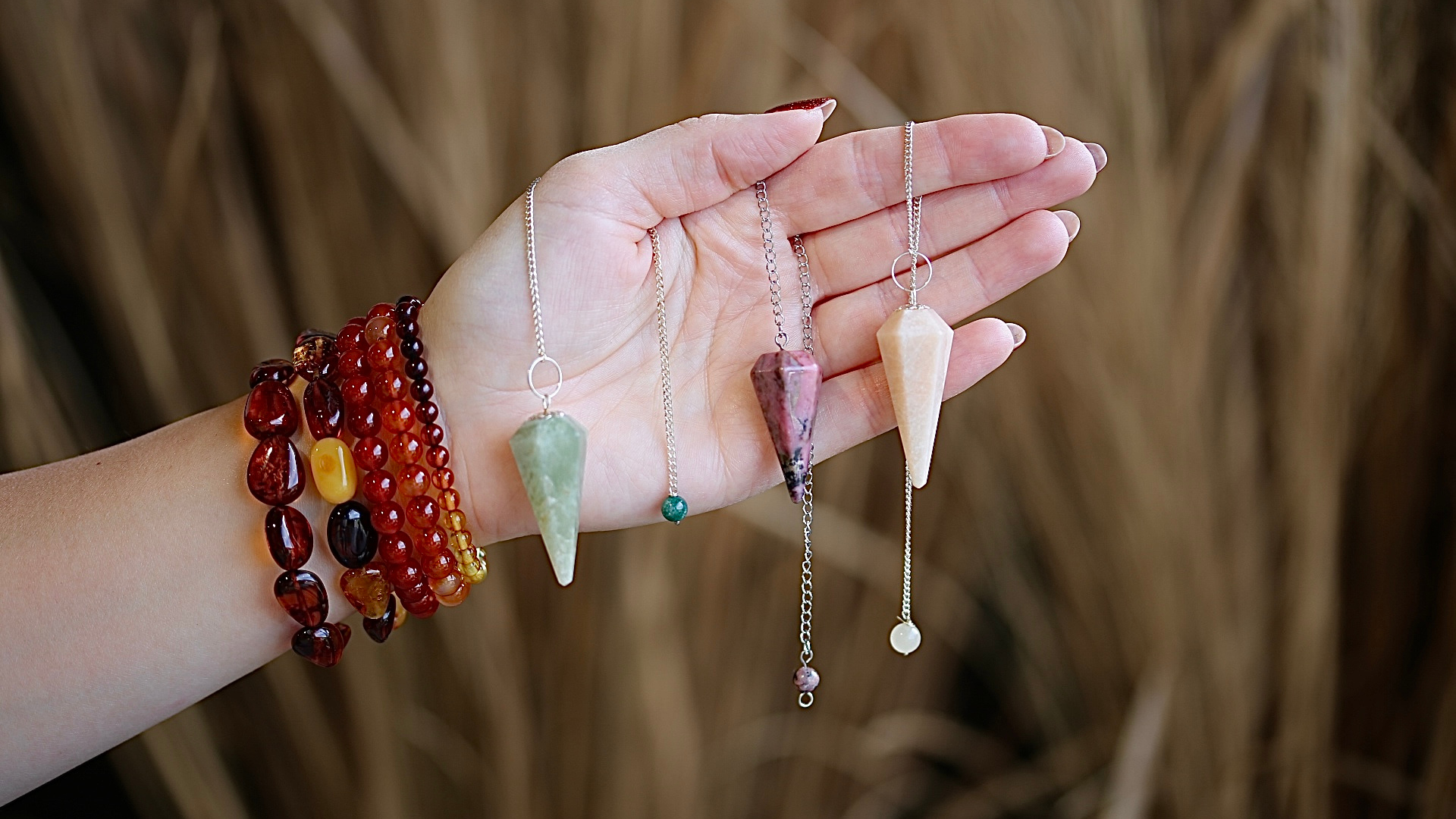 Hand waaraan diverse edelsteen pendels hangen. Om de pols barnsteen armbanden en carneool armbanden.